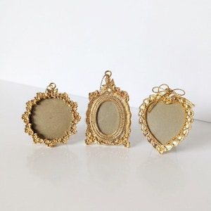 May include: Three small gold-toned picture frames with intricate decorative borders. The frames include a circular frame, a rectangular frame with rounded corners, and a heart-shaped frame, each featuring a small hanging loop at the top.