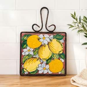 May include: A square ceramic tile featuring a hand-painted design of bright yellow lemons, green leaves, and white flowers on a reddish-brown background. The tile is set within a dark metal frame with a decorative scrolled handle at the top.
