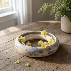 May include: A circular, cream-coloured ceramic ring vase featuring a hand-painted blue floral pattern. The hollow centre of the vase is decorated with several small, delicate yellow pansy flowers placed inside the basin and on the rim.
