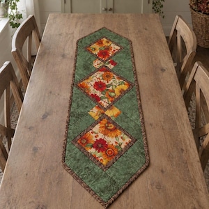 May include: A quilted table runner featuring a series of diamond-shaped panels with a floral pattern in shades of orange, red, and yellow, bordered by a textured sage green fabric and a dark brown patterned trim, laid out in a linear arrangement.