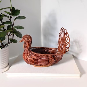 May include: A brown woven wicker basket shaped like a turkey. The basket features a rounded body with a hollow centre for storage, a detailed head with a beak and wattle, and a tall, fanned tail made of looped wicker strands.