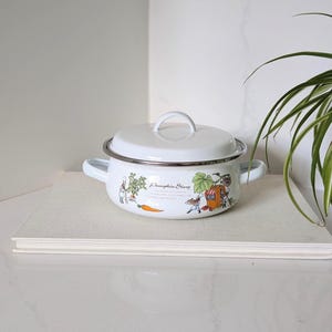 Peut inclure: Une marmite en émail blanc avec un couvercle et deux poignées latérales. L'extérieur présente une illustration colorée de petits personnages, une carotte et une structure en forme de citrouille avec le texte Pumpkin Story en écriture cursive.