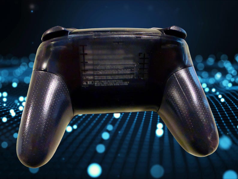 K&ouml;nnte beinhalten: Ein durchscheinend schwarzer kabelloser Videospiel-Controller von der R&uuml;ckseite gesehen. Das Ger&auml;t verf&uuml;gt &uuml;ber einen strukturierten Griff an beiden Seiten und ein zentrales Batteriefach mit schwach gepr&auml;gtem Text und sichtbaren Kennzeichnungen.