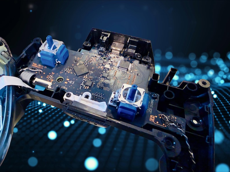 K&ouml;nnte beinhalten: Eine freiliegende interne Leiterplatte eines Videospiel-Controllers mit zwei blauen analogen Stick-Modulen, verschiedenen Mikrochips und integrierten elektronischen Komponenten, die auf einer dunkelgr&uuml;nen Platine in einem schwarzen Kunststoffgeh&auml;use montiert sind.