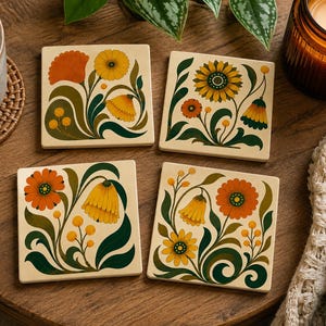 May include: A set of four square coasters featuring a cream-colored background with stylized floral illustrations in shades of orange, yellow, and dark green. Each coaster displays a unique arrangement of blooming flowers, curved stems, and leaves.