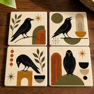May include: A set of four square coasters featuring cream-colored backgrounds with black crow illustrations. Each coaster displays geometric shapes, starbursts, and botanical elements in shades of olive green, burnt orange, and mustard yellow.