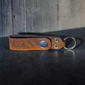 May include: A tan leather keychain featuring a laser-engraved bald eagle and American flag design. The strap is secured with a silver-toned metal snap button and two rivets, and it includes a circular silver-toned metal split ring for keys.