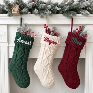 以下が含まれることがあります： ダークグリーン、クリーム、深紅のケーブルニット製クリスマスストッキング3足。各ストッキングの折り返し部分には、それぞれAmari、Mayra、Theoという名前が刺繍されています。ストッキングには人工の松の枝と赤いベリーが詰められています。