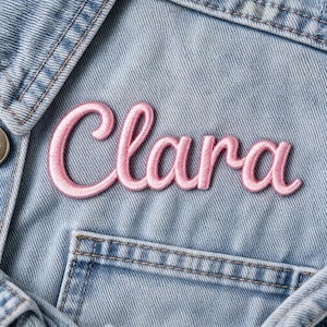 Op de afbeelding: Een roze geborduurde patch met de naam "Clara" in een sierlijk handschrift. De patch is genaaid op een lichtblauwe spijkerjas, met de naam boven een zak. De borduursels zijn gedetailleerd en de roze kleur is levendig.