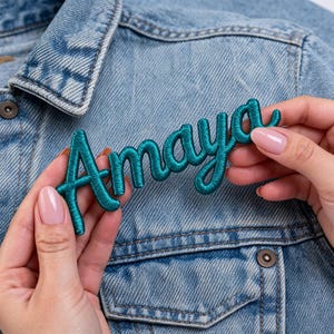 Op de afbeelding: Teal geborduurde patch met de naam "Amaya" in een sierlijk lettertype. De patch wordt getoond tegen een lichtblauwe spijkerjas. De teal kleur van de patch contrasteert met het blauw van de jas.