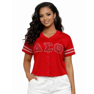 May include: Red baseball jersey with white stripes on the sleeves. The jersey features the Greek letters ΔΣΟ in a sparkling, embellished design. The jersey has a button-up front and is paired with white shorts.