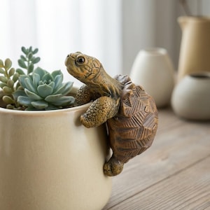 Puede incluir: Un adorno de tortuga de cerámica marrón y beige, agarrado al borde de una maceta beige llena de suculentas. La maceta está sobre una superficie de madera, con otros jarrones de cerámica en el fondo.