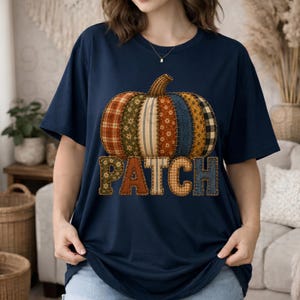 Op de afbeelding: Een marineblauw t-shirt met korte mouwen en een grote pompoenafbeelding gemaakt van verschillende stoffen patronen, waaronder ruitjes, bloemen en denim. Onder de pompoen staat het woord PATCH in grote, blokvormige letters met textuur.