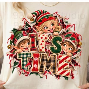 Op de afbeelding: Een crèmekleurig T-shirt met een afbeelding van drie cartoonelfjes in rood-groene mutsen die door een gescheurd stofeffect kijken. De tekst IT'S TIME wordt weergegeven in grote, getextureerde letters met diverse patronen zoals ruiten en sneeuwvlokken.