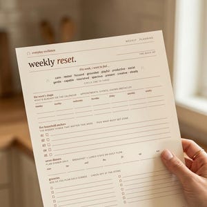 May include: A beige weekly planner with the text "weekly reset" and sections for planning. The planner includes prompts for goals, appointments, and groceries. The planner is held by a person.