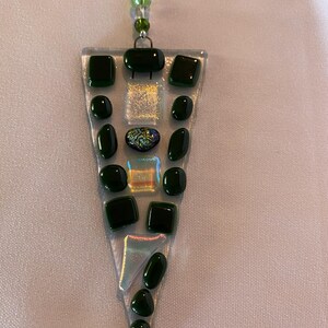 May include: A clear glass pendant with a triangular shape, adorned with dark green glass pieces in various shapes. The pendant is suspended by a beaded strand with green beads. The design includes square, oval, and rectangular glass elements.