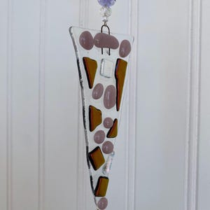 May include: A triangular fused glass suncatcher featuring a clear base decorated with various shapes of opaque mauve and translucent amber glass pieces. The ornament hangs from a metal loop adorned with a single faceted light purple glass bead.