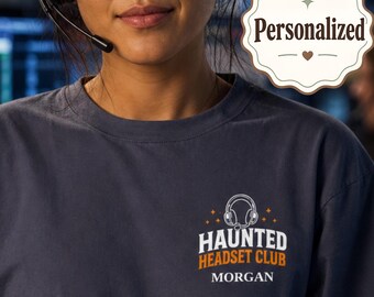 May include: A dark gray short-sleeve t-shirt featuring a graphic on the chest with an illustration of a headset, the text "HAUNTED HEADSET CLUB" in white and orange, and the name "MORGAN" printed below in white. A "Personalized" badge is visible.