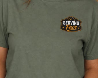 May include: An olive green short-sleeved t-shirt featuring a black and orange graphic on the left chest. The graphic is a decorative badge shape containing the text "SERVING Face" in white and orange lettering, accented with small stars.