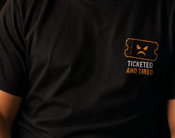 May include: A black short-sleeved t-shirt featuring a small graphic on the left chest. The graphic depicts an orange ticket icon with a frowning face and the text "TICKETED AND TIRED" printed in orange and white lettering below the icon.