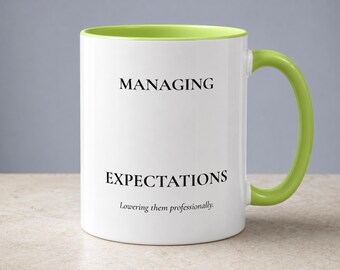 May include: A white ceramic mug with a lime green interior and handle. The front features black text that reads: MANAGING EXPECTATIONS, Lowering them professionally. The mug has a cylindrical body and a curved handle on the right side.