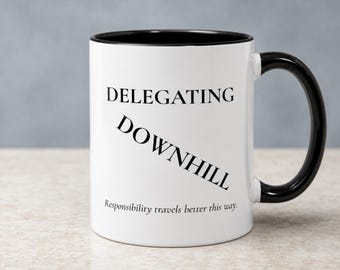 May include: A white ceramic mug with a black interior and a black handle. The front features the black text DELEGATING in a serif font, followed by the word DOWNHILL printed diagonally, and the phrase Responsibility travels better this way.