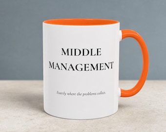 May include: A white ceramic mug with a bright orange interior and handle. The front features the black text "MIDDLE MANAGEMENT" in a bold serif font, followed by the smaller text "Exactly where the problems collect." below it.
