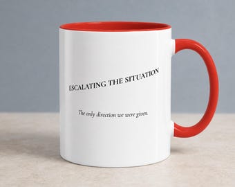 May include: A white ceramic mug with a bright red interior and handle. The front features the black text ESCALATING THE SITUATION in all caps, followed by the smaller black text The only direction we were given. The mug has a cylindrical shape.