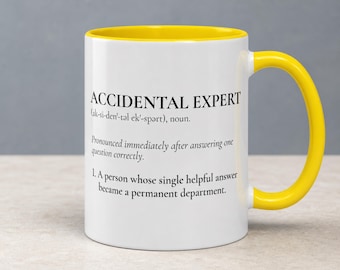 May include: A white ceramic mug with a bright yellow interior and handle. The front features black printed text that reads: ACCIDENTAL EXPERT (ak-si-den-təl ek-spərt), noun. Pronounced immediately after answering one question correctly. 1. A person whose single helpful answer became a permanent department.
