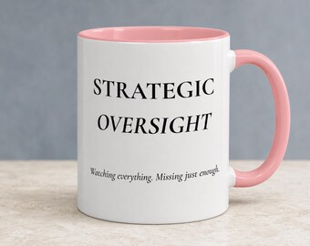 May include: A white ceramic mug with a soft pink interior and handle. The front features the black text STRATEGIC OVERSIGHT in a serif font, followed by the smaller black text Watching everything. Missing just enough. below it.