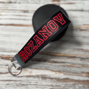May include: A black leather wristlet keychain featuring the name ROZANOV printed in bold red varsity-style lettering. The strap is attached to a silver-toned metal clasp and a circular split ring, designed for holding keys or other small items.