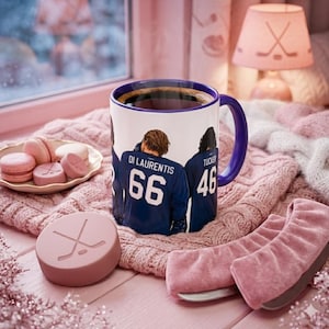 Puede incluir: Una taza de cerámica blanca con borde y asa azul oscuro, que presenta un gráfico de dos jugadores de hockey con camisetas azul oscuro con los nombres DI LAURENTIS 66 y TUCKER 46 impresos en la parte posterior, llena de café negro.