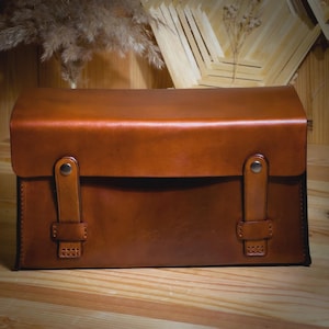 May include: A brown leather satchel with a rectangular shape and a curved top. The bag features two leather straps with metal closures and is set on a wooden surface. The leather has a glossy finish.