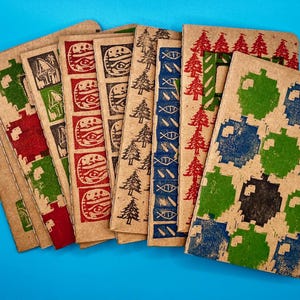 May include: A collection of kraft paper notebooks with various block-printed designs in red, green, blue, and black. The designs include trees, abstract shapes, and burger-like icons. The notebooks are arranged on a bright blue surface.
