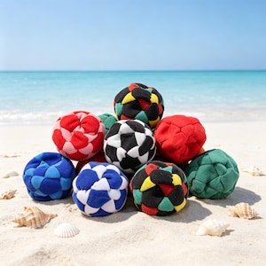 May include: A collection of colorful, patterned fabric balls, including red, blue, green, black, and white, are arranged on a sandy beach with seashells. The balls have geometric designs and are likely used for games or decorative purposes.