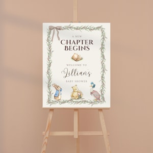 May include: A rectangular sign on a wooden easel featuring a green vine border with a brown ribbon. The text reads A NEW CHAPTER BEGINS, WELCOME TO, Jillian's, BABY SHOWER. Illustrations include an open book, a rabbit, a bear, and a duck.