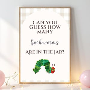 May include: A framed print with the text "CAN YOU GUESS HOW MANY book worms ARE IN THE JAR?" features a green caterpillar illustration. The print is in a wooden frame, with a light beige and white checkered pattern at the top. Two pink balloons are at the bottom.