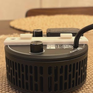 May include: A black A360 light fixture with a white mounting bracket and black knobs. The fixture has a cylindrical shape with vertical vents and a black power cord. The fixture is sitting on a woven surface.