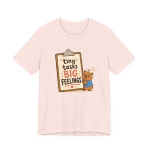 May include: A pale pink t-shirt with a cartoon graphic. The design features a clipboard with the words "tiny tasks BIG FEELINGS" and a bear character dressed as a nurse. The bear is holding the clipboard, with a small heart below.