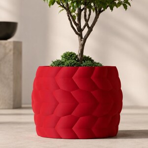 May include: A vibrant red cylindrical planter featuring a repeating embossed chevron or leaf-like pattern across its exterior surface. A small green tree with a thin brown trunk and dense foliage is planted inside the container.
