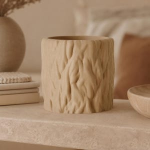 Pode incluir: Um vaso cilíndrico bege com um exterior texturizado que imita a aparência da casca de uma árvore. O recipiente tem uma abertura larga no topo e um acabamento mate consistente em toda a superfície, exibindo um padrão orgânico detalhado.