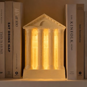 May include: A cream-colored, temple-shaped lamp featuring four fluted columns, a triangular pediment, and a rectangular base. The structure is illuminated from within, casting a warm, soft glow through the columns and the interior space.
