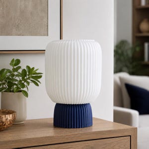 May include: A modern white lamp with vertical ribbed detailing sits atop a navy blue base. The lamp is cylindrical with a rounded top. The lamp is on a wooden surface, with a plant and framed artwork in the background.