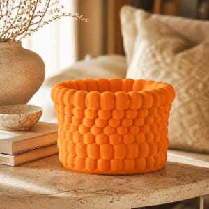 May include: A bright orange cylindrical container featuring a textured exterior composed of numerous small, rounded, bubble-like protrusions arranged in a repeating honeycomb pattern. The object has a wide, open top and a flat, circular base.