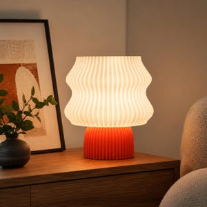 May include: A modern table lamp with a ribbed, orange base and a wavy, off-white shade. The lamp is turned on, emitting a warm glow. The lamp sits on a wooden nightstand, next to a framed artwork and a small vase with greenery.