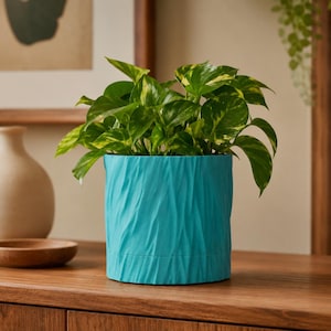 May include: A vibrant turquoise plant pot with a textured, rippled surface finish. The cylindrical container holds a lush pothos plant featuring heart-shaped green leaves with yellow variegation, creating a striking contrast against the bright blue.