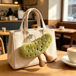 Op de afbeelding: Een crèmekleurige draagtas met een groene, pluche augurk sleutelhanger. De augurk heeft een lachend gezicht en bruine benen. De tas heeft twee handvatten en een zilveren sleutelhanger. De achtergrond toont een café met een kopje koffie.
