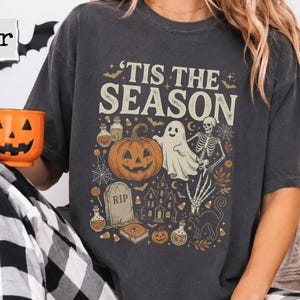 May include: A charcoal gray short-sleeve t-shirt featuring the text 'TIS THE SEASON' above a graphic illustration of a pumpkin, a ghost, a skeleton, a tombstone, a haunted house, and various potion bottles in orange, white, and black colors.
