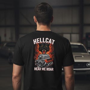May include: A black short-sleeve t-shirt featuring a graphic on the back. The design displays a snarling black panther head above a detailed silver engine block, surrounded by orange and red flames, with the text HELLCAT above and HEAR ME ROAR below.