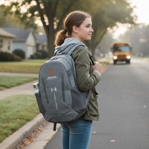 Peut inclure: Un sac à dos gris Under Armour avec une poche latérale en filet contenant une bouteille d'eau argentée. Le sac présente un petit logo circulaire orange au-dessus du texte Claude et le logo Under Armour imprimé en blanc sur le panneau avant.